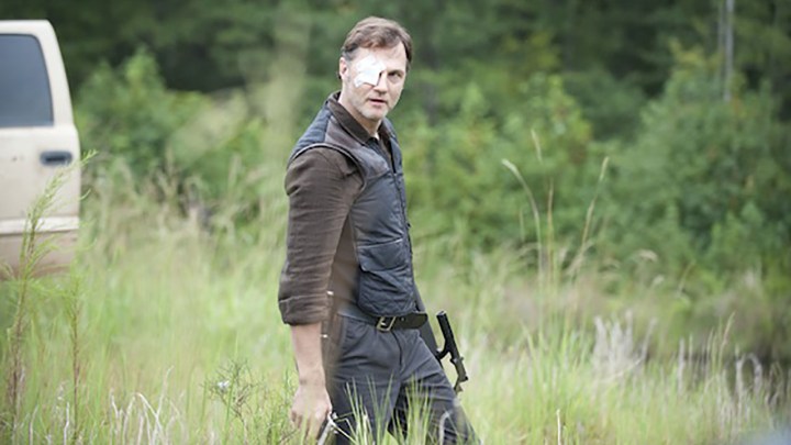 The Governor walking alone in the woods, holding a gun in a scene from The Walking Dead.