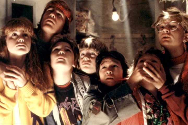 The cast of the Goonies is standing in a basement, dimly lit staring off into the distance.