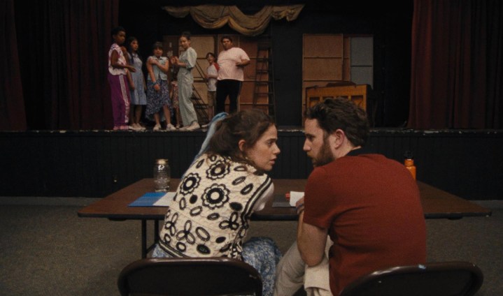 Two adults sit at a table as kids stand on a stage in Theater Camp.