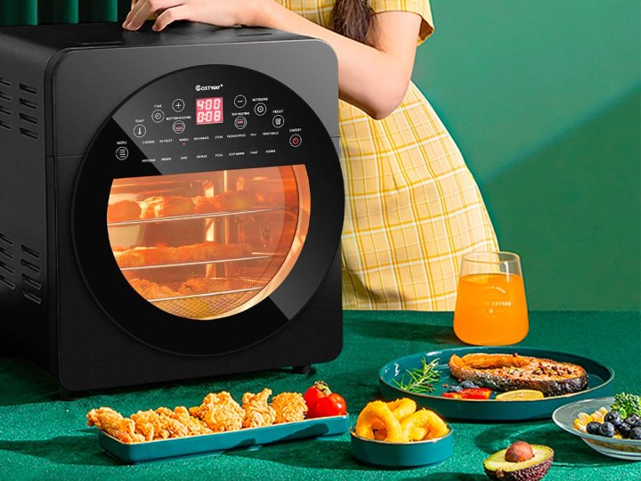 The Costway 16-in-1 15-quart air fryer oven on a table with plates of food spread out around it.