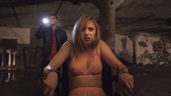 Maika Monroe bound to a chair with a man shining a flashlight behind her in It Follows.