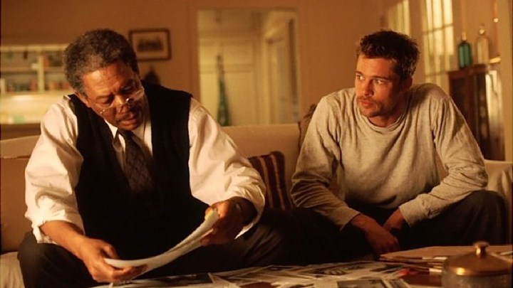 Morgan Freeman and Brad Pitt sit together on a couch looking at papers in a scene from Seven.