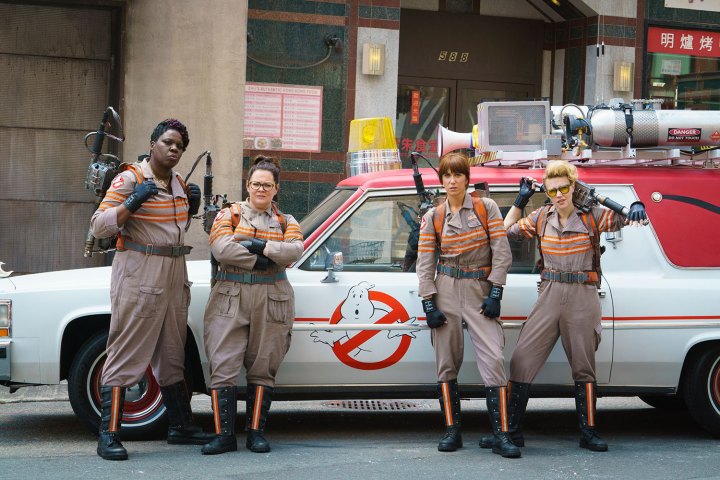 Four female ghostbusters stand in front of a car.