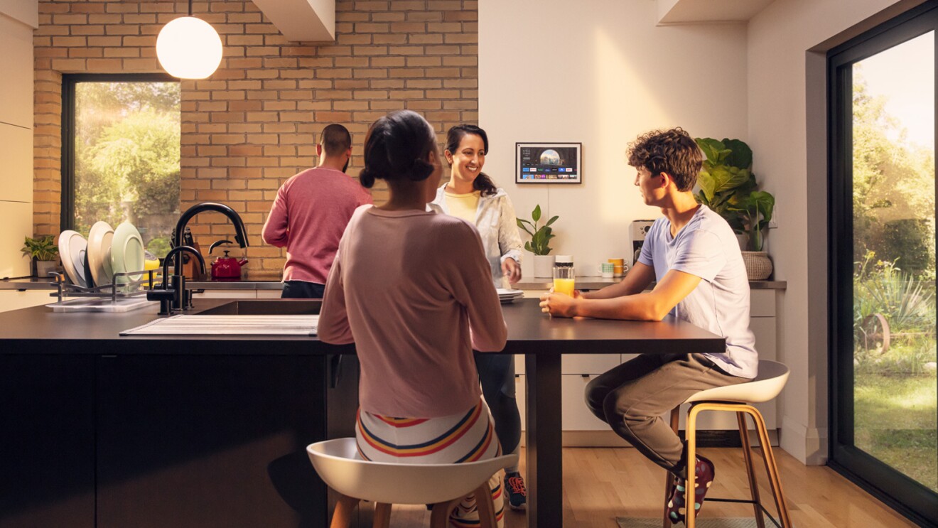 An image of four people interacting in a kitchen with an Echo Show 15 mounted to the wall in the background. 